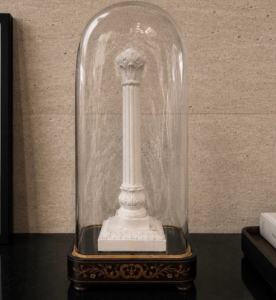 White marble column sculpture under a glass dome on a dark surface with a textured beige wall background. dome on a wooden surface. Antique French glass dome large oval