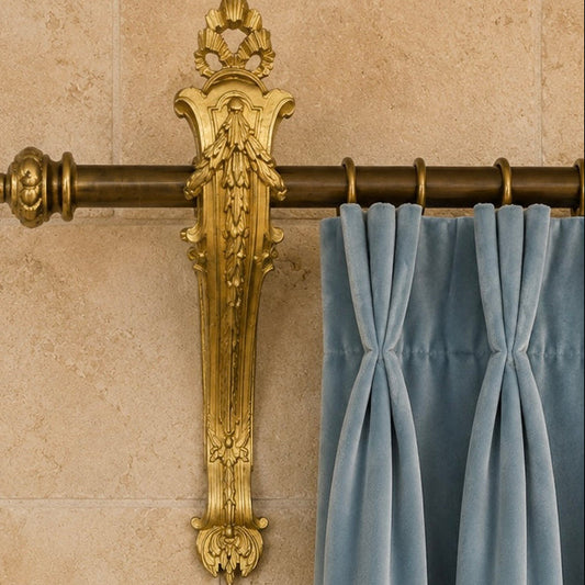 Decorative antique french gold curtain rod  bracketwith intricate design against a beige wall with blue curtains.