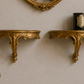 Two ornate gold wall-mounted console shelves with carved details, displayed on a textured beige wall beneath an oval gold mirror. A small black candle sits on the right shelf, and part of a matching upholstered chair is visible in the lower left corner.